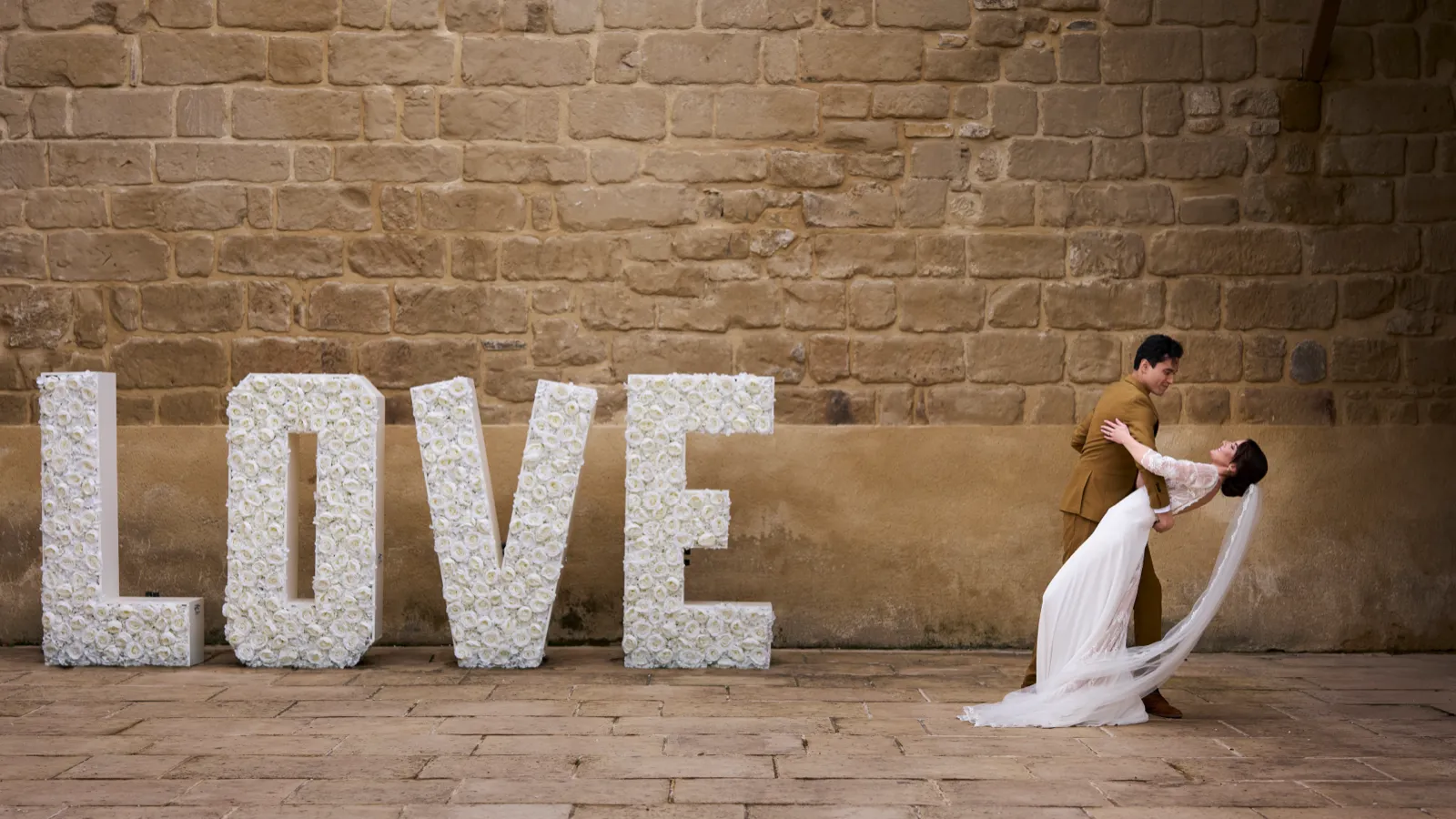 Couple de mariés enlacé devant des lettres florales géantes formant le mot LOVE, contre un mur de pierre